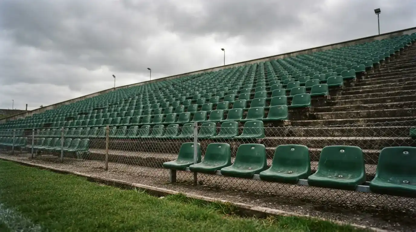 Rzędy pustych plastikowych krzesełek małego stadionu widziane z poziomu murawy pod pochmurnym niebem.