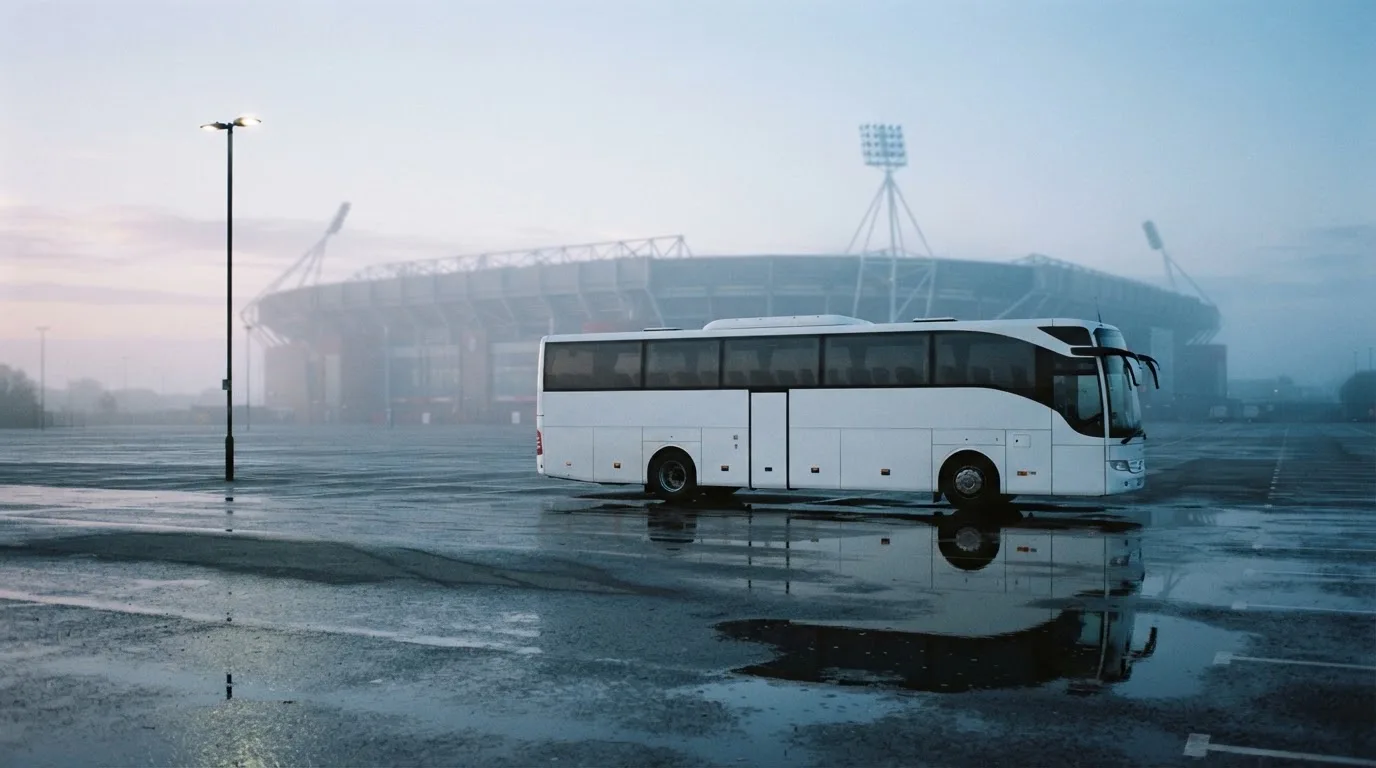 Autokar stoi na parkingu przed stadionem o świcie, rozmyte kontury trybun w tle, pusty plac i mokry asfalt.